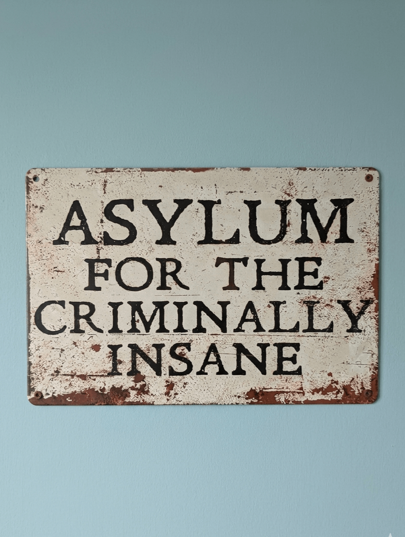 Landscape-oriented white metal wall sign with the bold black text 'ASYLUM FOR THE CRIMINALLY INSANE' featuring heavy faux-rust and distressed textures on a light blue wall.