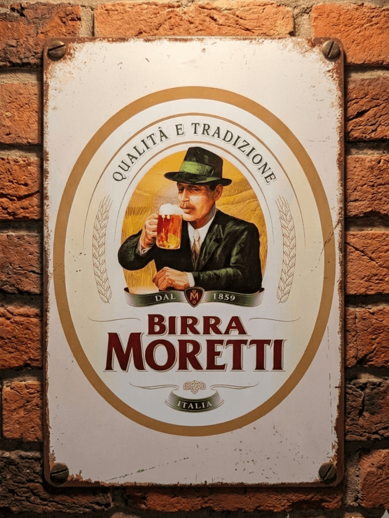 Vertical vintage-style metal wall sign for Birra Moretti, featuring the iconic illustration of a man with a moustache in a green hat drinking a beer, centered in an oval frame with 'Qualità e Tradizione' text on a distressed white background, mounted on a brick wall.