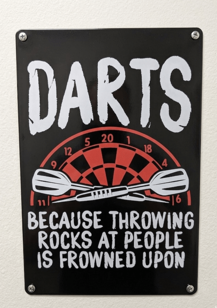 Vertical black metal wall sign with the text 'DARTS BECAUSE THROWING ROCKS AT PEOPLE IS FROWNED UPON' in white distressed typography, featuring an illustration of a red and black dartboard with two crossed darts.