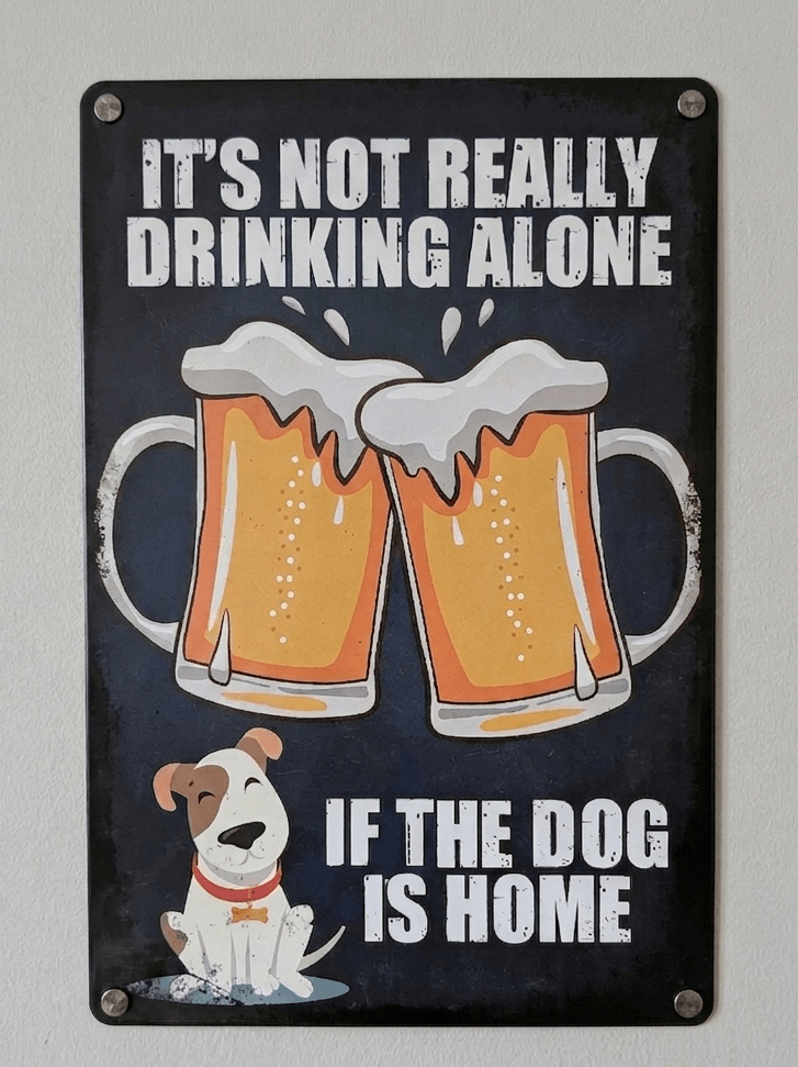Vertical vintage-style metal wall sign with the text 'IT'S NOT REALLY DRINKING ALONE IF THE DOG IS HOME' in bold white letters, featuring an illustration of two clinking beer mugs and a small, happy dog at the bottom on a distressed dark blue background.
