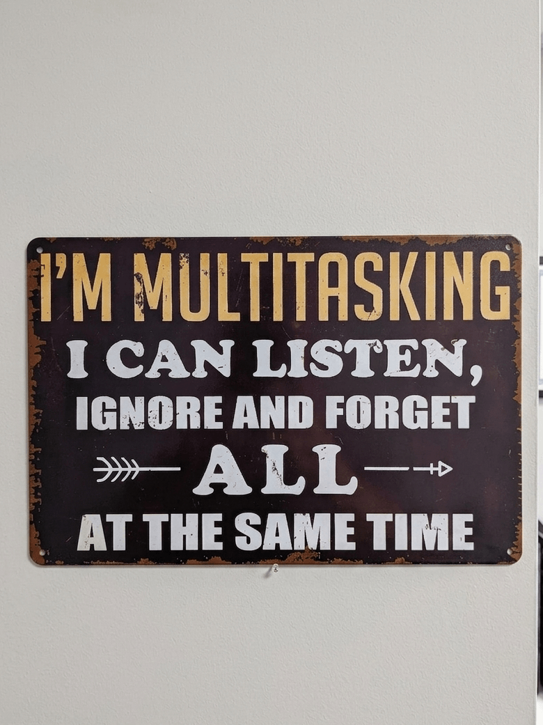 Horizontal vintage-style metal wall sign with the text 'I'M MULTITASKING I CAN LISTEN, IGNORE AND FORGET ALL AT THE SAME TIME' in yellow and white typography on a dark, distressed background with rusted edges