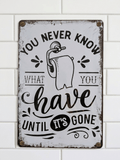 Vertical vintage-style metal wall sign for a bathroom featuring a toilet roll illustration and the text 'YOU NEVER KNOW WHAT YOU HAVE UNTIL IT'S GONE' in black typography on a distressed white background, mounted on a white subway tile wall.