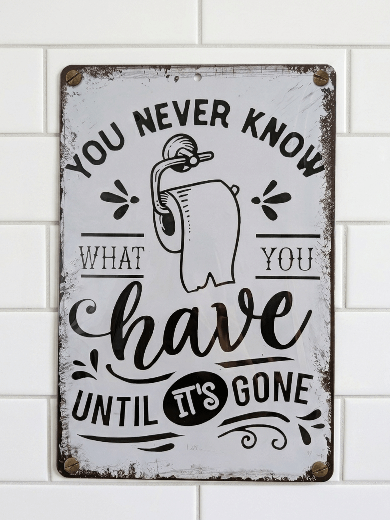 Vertical vintage-style metal wall sign for a bathroom featuring a toilet roll illustration and the text 'YOU NEVER KNOW WHAT YOU HAVE UNTIL IT'S GONE' in black typography on a distressed white background, mounted on a white subway tile wall.