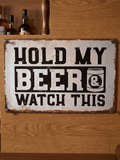 Horizontal vintage-style metal wall sign mounted on wood reading 'HOLD MY BEER & WATCH THIS' in bold black block lettering with a small beer can graphic on a distressed off-white background with heavily rusted borders.