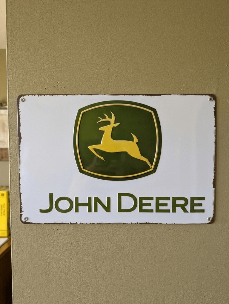 Classic John Deere logo metal wall sign featuring the yellow leaping deer in a green shield on a clean white background with a slightly distressed border