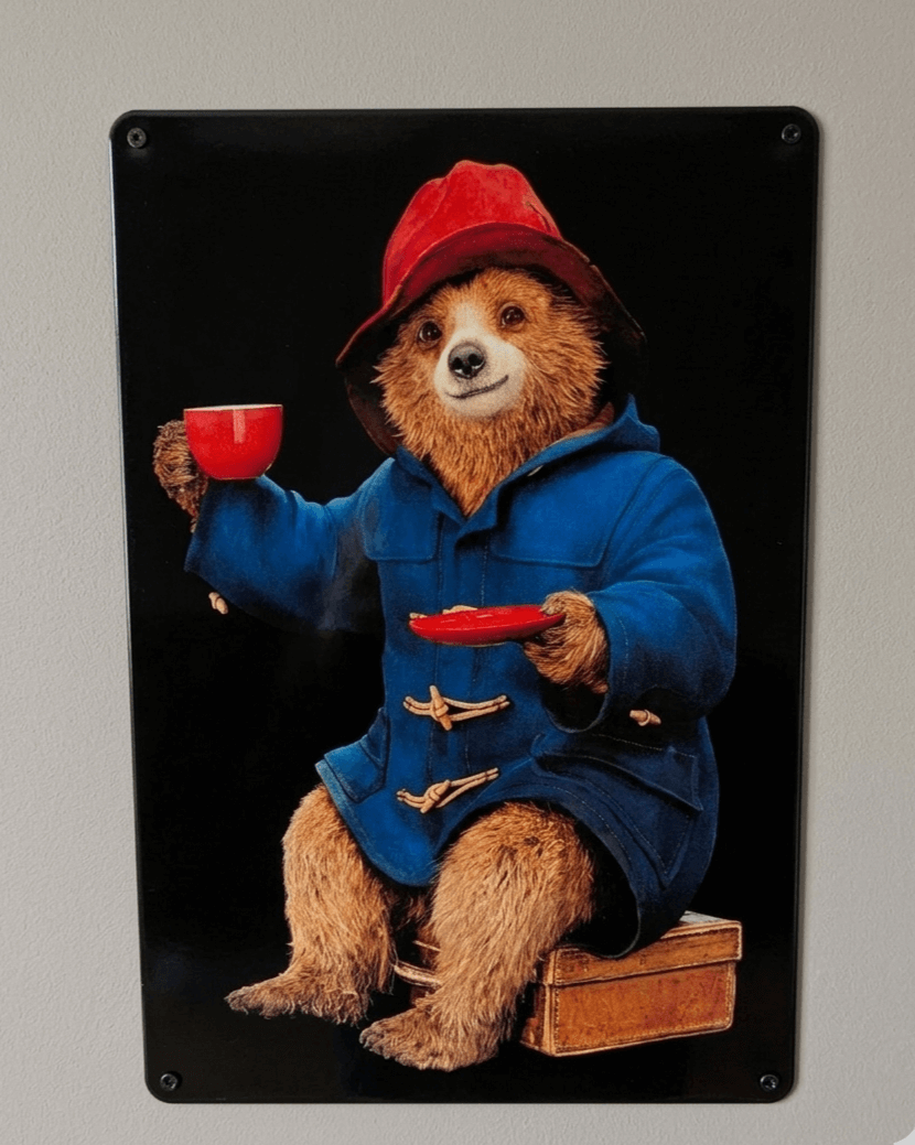 Vibrant metal wall sign of Paddington Bear in his blue duffle coat and red hat, sitting on his suitcase and holding a red cup of tea against a black background