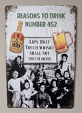 Vertical vintage-style metal wall sign reading 'REASONS TO DRINK NUMBER 452' at the top, featuring a vintage black and white photo of an unamused group of women holding a sign that says 'LIPS THAT TOUCH WHISKY SHALL NOT TOUCH OURS', flanked by color illustrations of a liquor bottle and a frothy beer mug on a distressed white background with heavily rusted borders.