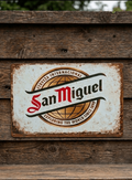 Landscape-oriented metal wall sign for San Miguel beer, featuring the 'Cerveza Internacional' globe logo and 'Exploring the world since 1890' text on a heavily distressed white background.