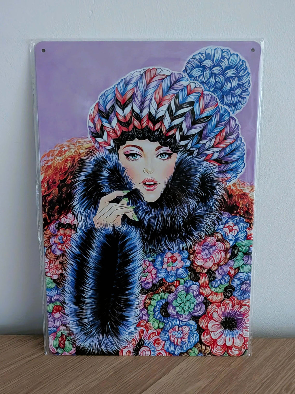 Vertical glossy metal wall sign in protective plastic wrap featuring Sunny Gu's fashion illustration 'Keep Warm', showing a chic woman in a thick patterned knit hat, large dark fur collar, and vibrant colorful floral patterned coat against a soft purple background.