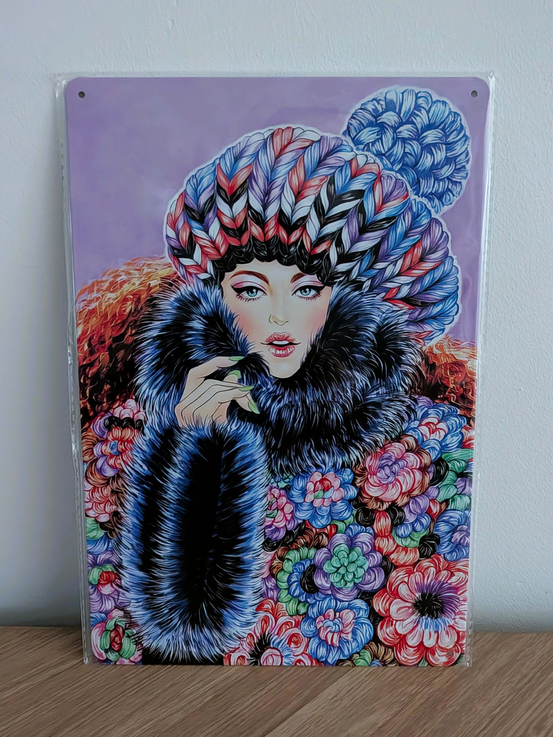 Vertical glossy metal wall sign in protective plastic wrap featuring Sunny Gu's fashion illustration 'Keep Warm', showing a chic woman in a thick patterned knit hat, large dark fur collar, and vibrant colorful floral patterned coat against a soft purple background.