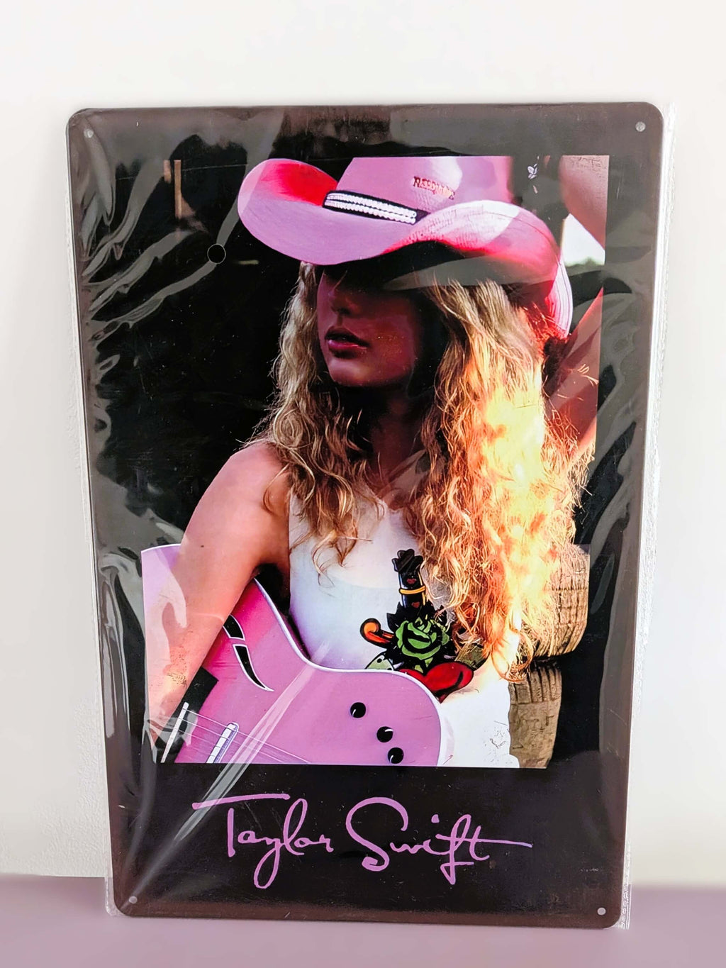 Vertical glossy metal wall sign for Taylor Swift in protective plastic wrap, featuring a retro photographic print of Taylor with curly blonde hair, a pink cowboy hat, and a pink acoustic guitar over a black border with her signature logo printed in pink at the bottom.