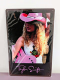 Vertical glossy metal wall sign for Taylor Swift in protective plastic wrap, featuring a retro photographic print of Taylor with curly blonde hair, a pink cowboy hat, and a pink acoustic guitar over a black border with her signature logo printed in pink at the bottom.