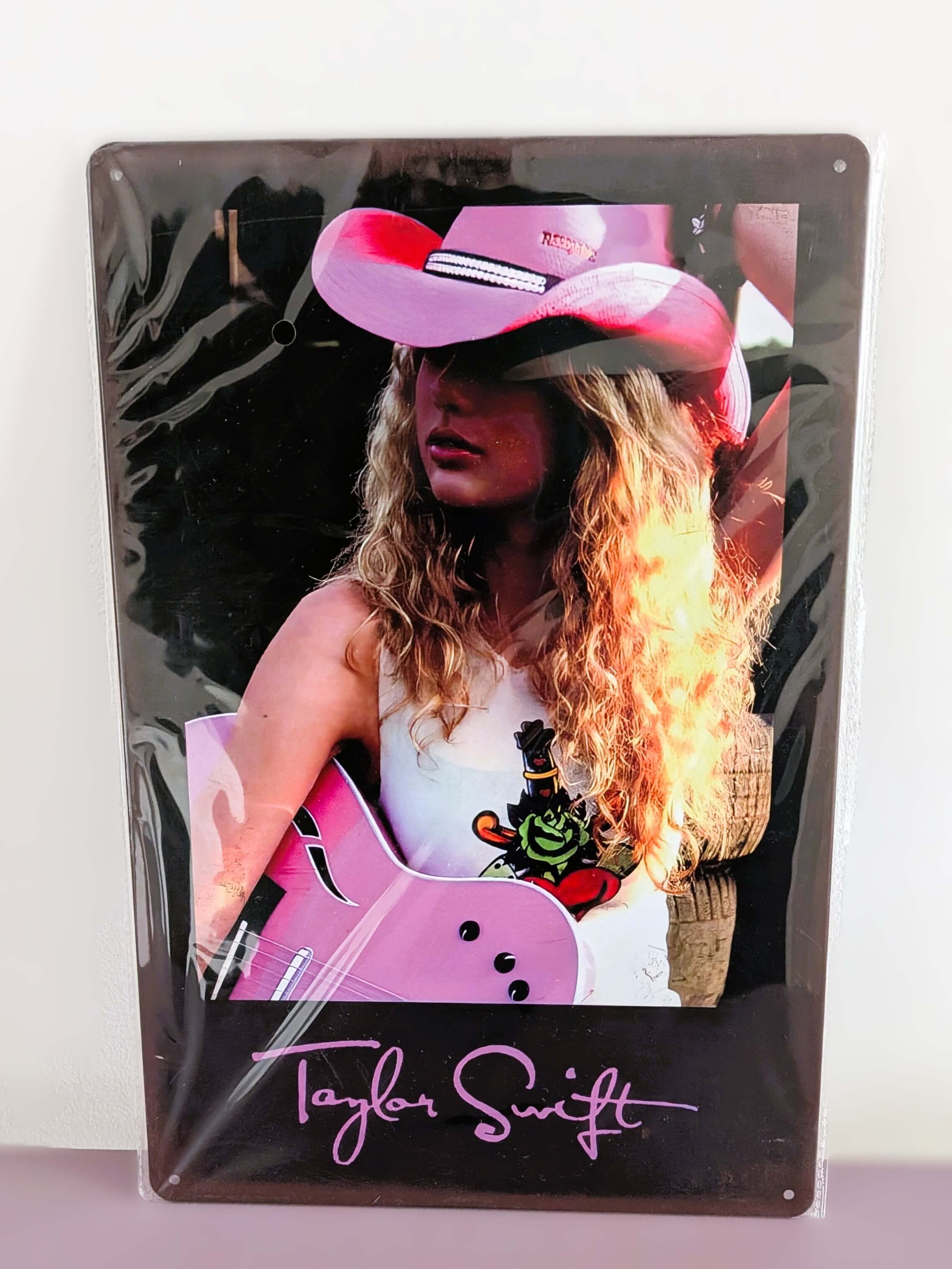 Vertical glossy metal wall sign for Taylor Swift in protective plastic wrap, featuring a retro photographic print of Taylor with curly blonde hair, a pink cowboy hat, and a pink acoustic guitar over a black border with her signature logo printed in pink at the bottom.