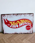 Horizontal vintage-style metal wall sign for Hot Wheels in a protective plastic wrap, featuring the classic red, yellow, and white flame logo with the Mattel seal on a heavily distressed white background with rusted borders.