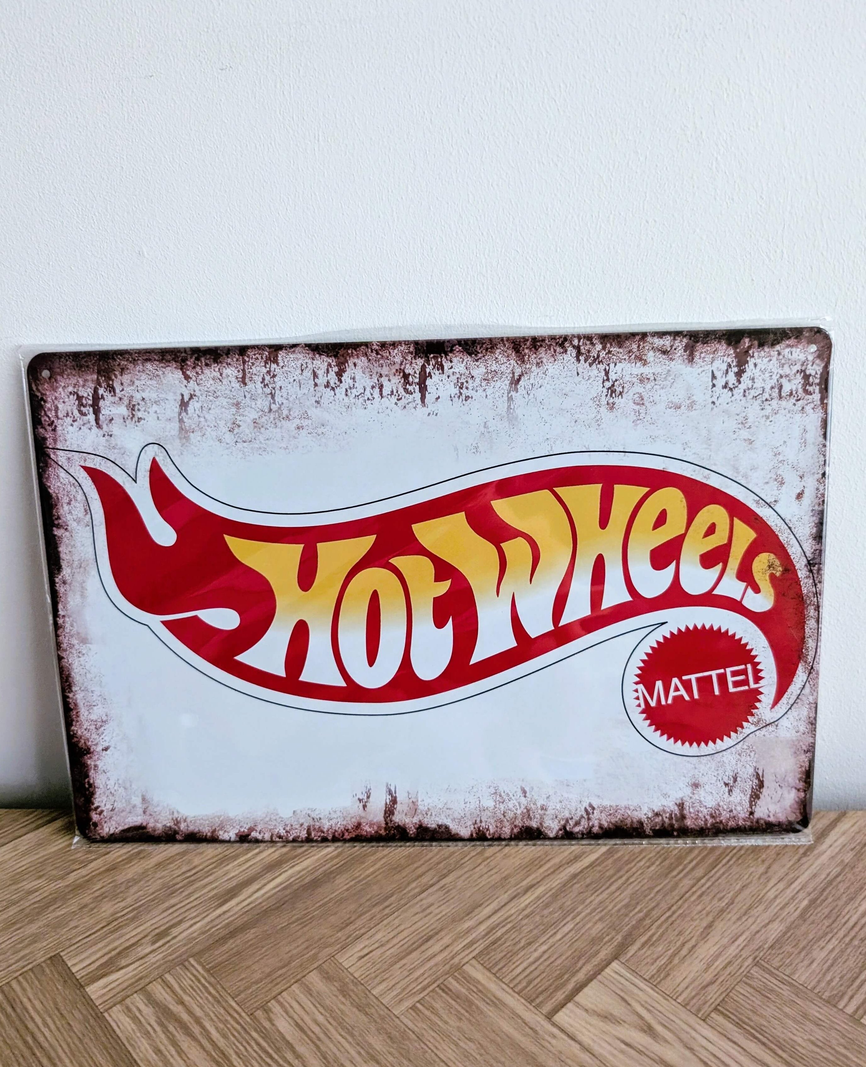 Horizontal vintage-style metal wall sign for Hot Wheels in a protective plastic wrap, featuring the classic red, yellow, and white flame logo with the Mattel seal on a heavily distressed white background with rusted borders.
