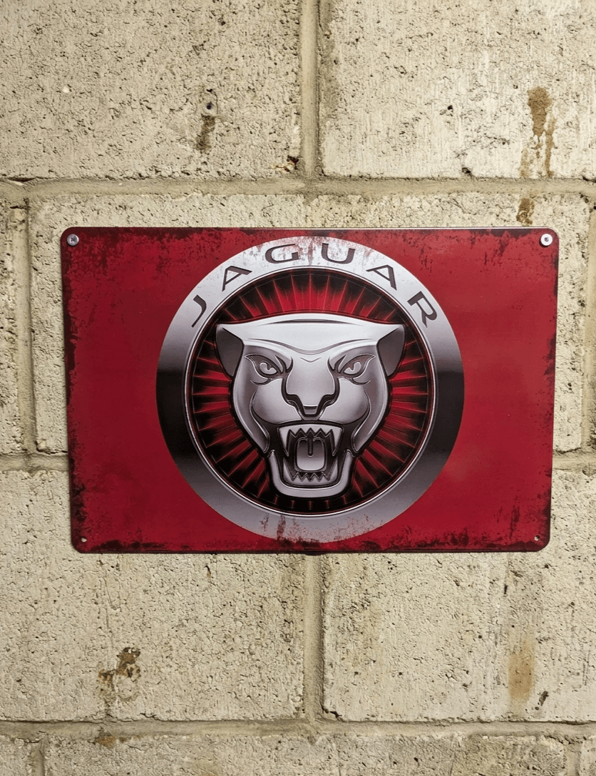 Horizontal vintage-style metal wall sign for Jaguar, featuring the silver Growler head logo centered on a vibrant, distressed red background, mounted on a light-colored brick wall.