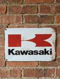 Vintage-style landscape metal wall sign for Kawasaki motorcycles, featuring the classic red 'Flying K' logo and black Kawasaki block lettering on a distressed white background with rusted edges.