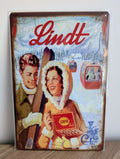 Vertical vintage-style metal wall sign for Lindt chocolate in protective plastic wrap, featuring a retro illustration of a smiling couple with skis and an open box of Lindor truffles against a snowy Alpine landscape with cable cars and heavily rusted borders.