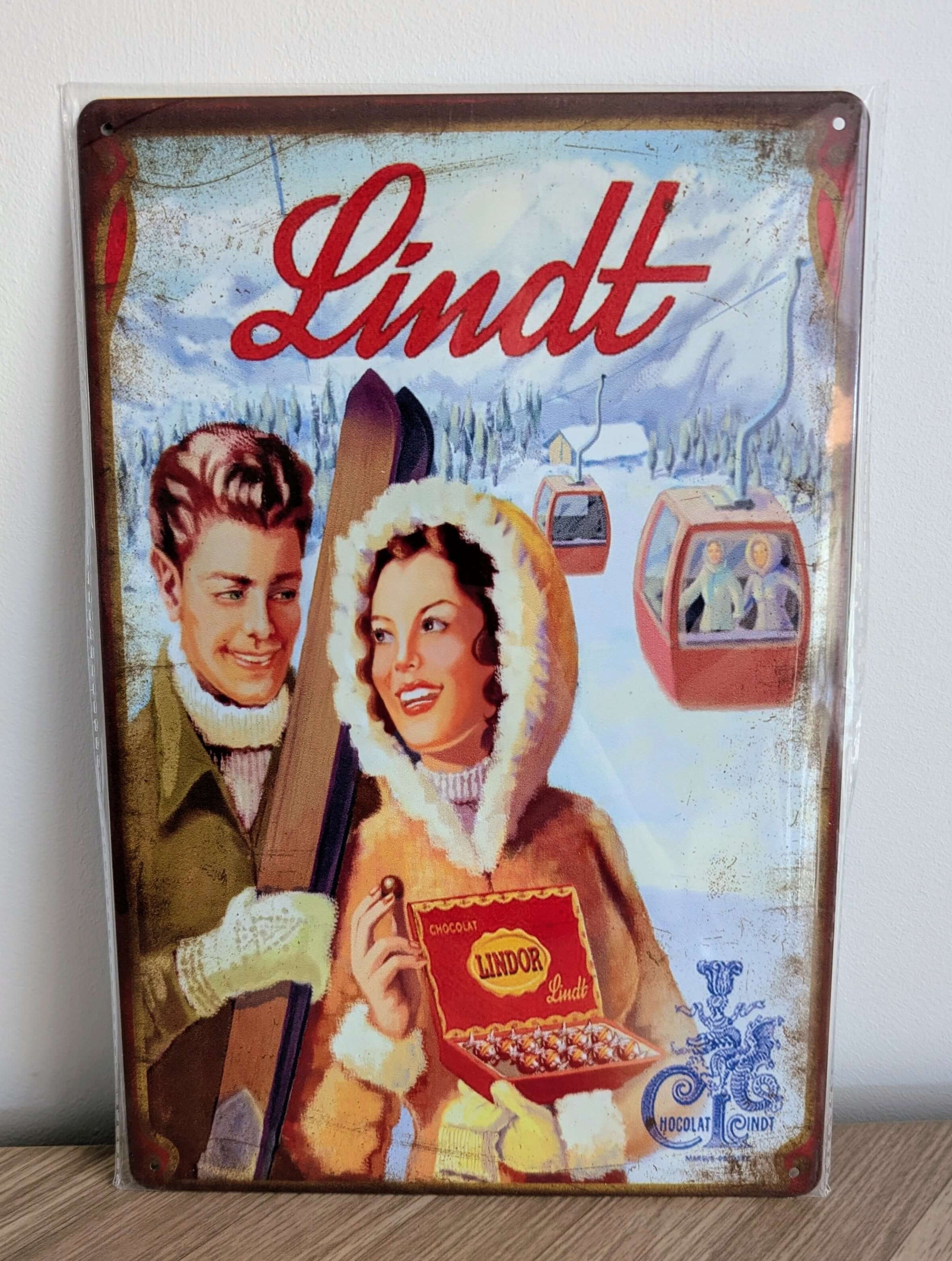 Vertical vintage-style metal wall sign for Lindt chocolate in protective plastic wrap, featuring a retro illustration of a smiling couple with skis and an open box of Lindor truffles against a snowy Alpine landscape with cable cars and heavily rusted borders.