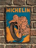 Vintage-style metal wall sign featuring the Michelin Man riding a motorcycle at high speed with orange and blue action graphics and a distressed rusted border