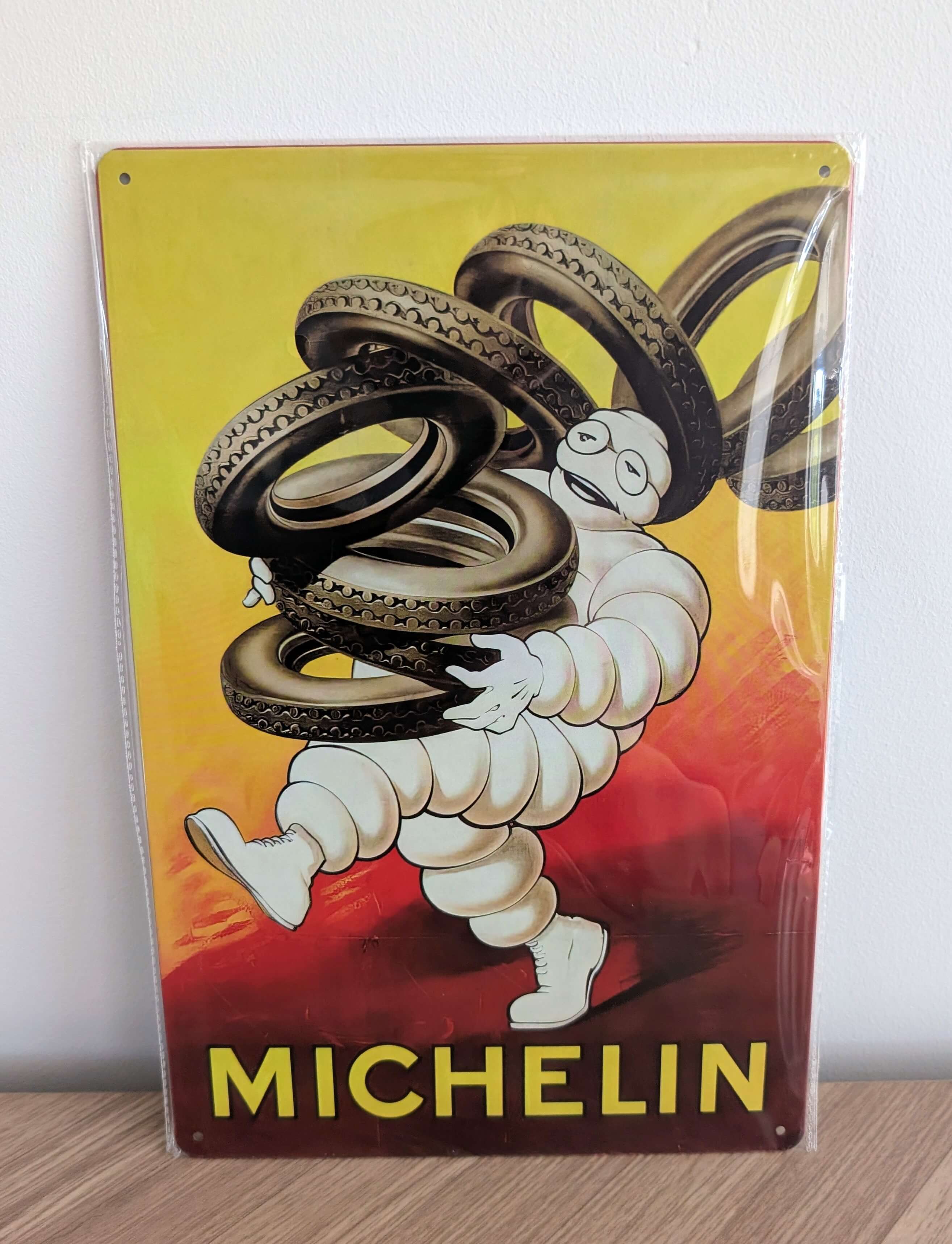 Vertical vintage-style metal wall sign for Michelin Tyres in protective plastic wrap, featuring the classic Michelin Man character playfully carrying a large stack of black tyres over his shoulder against a vibrant yellow and red gradient background with bold yellow text.
