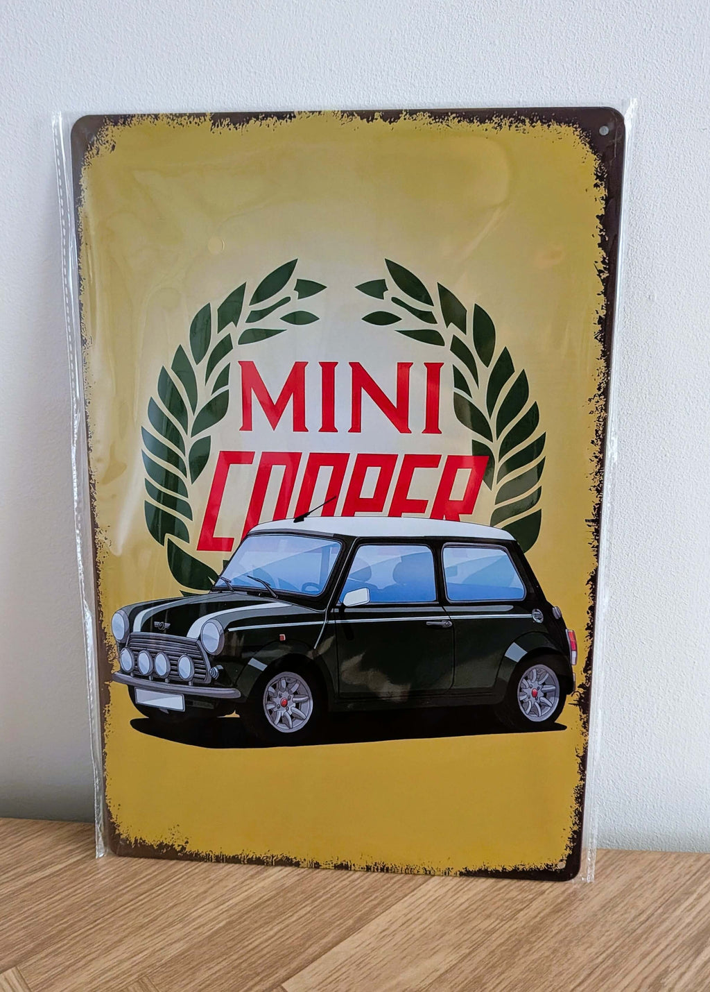 Vertical vintage-style metal wall sign for Mini Cooper in protective plastic wrap, featuring an illustration of a classic dark green Mini with a white roof in front of a green laurel wreath logo on a distressed yellow background with heavily rusted borders.