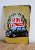 Vertical vintage-style metal wall sign for Mini Cooper in protective plastic wrap, featuring an illustration of a classic dark green Mini with a white roof in front of a green laurel wreath logo on a distressed yellow background with heavily rusted borders.