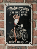 Vintage-style vertical metal wall sign featuring a pin-up woman posing with a classic motorcycle and the humorous text 'Motorcycles Are Like Wives: If It Ain’t Yours, Don’t Touch It' on a distressed black and cream background.