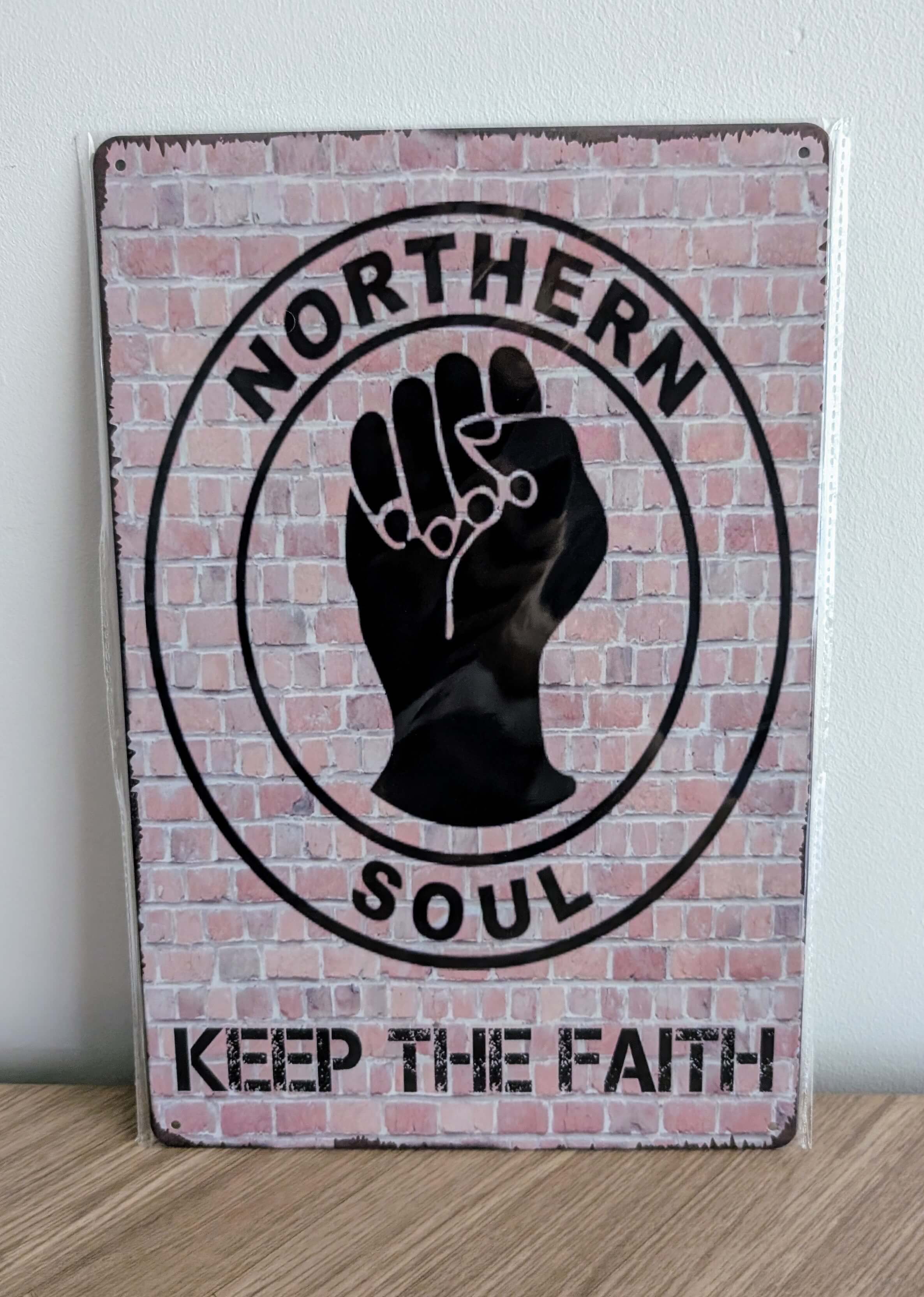 Vertical vintage-style metal wall sign for Northern Soul in protective plastic wrap, featuring the iconic black clenched fist insignia over a rustic pink brick wall background with 'KEEP THE FAITH' text at the bottom and distressed rusted edges.