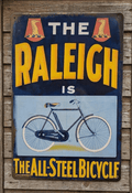 Vintage-style navy and yellow metal wall sign for Raleigh Bicycles featuring a classic bicycle illustration and the text The All-Steel Bicycle