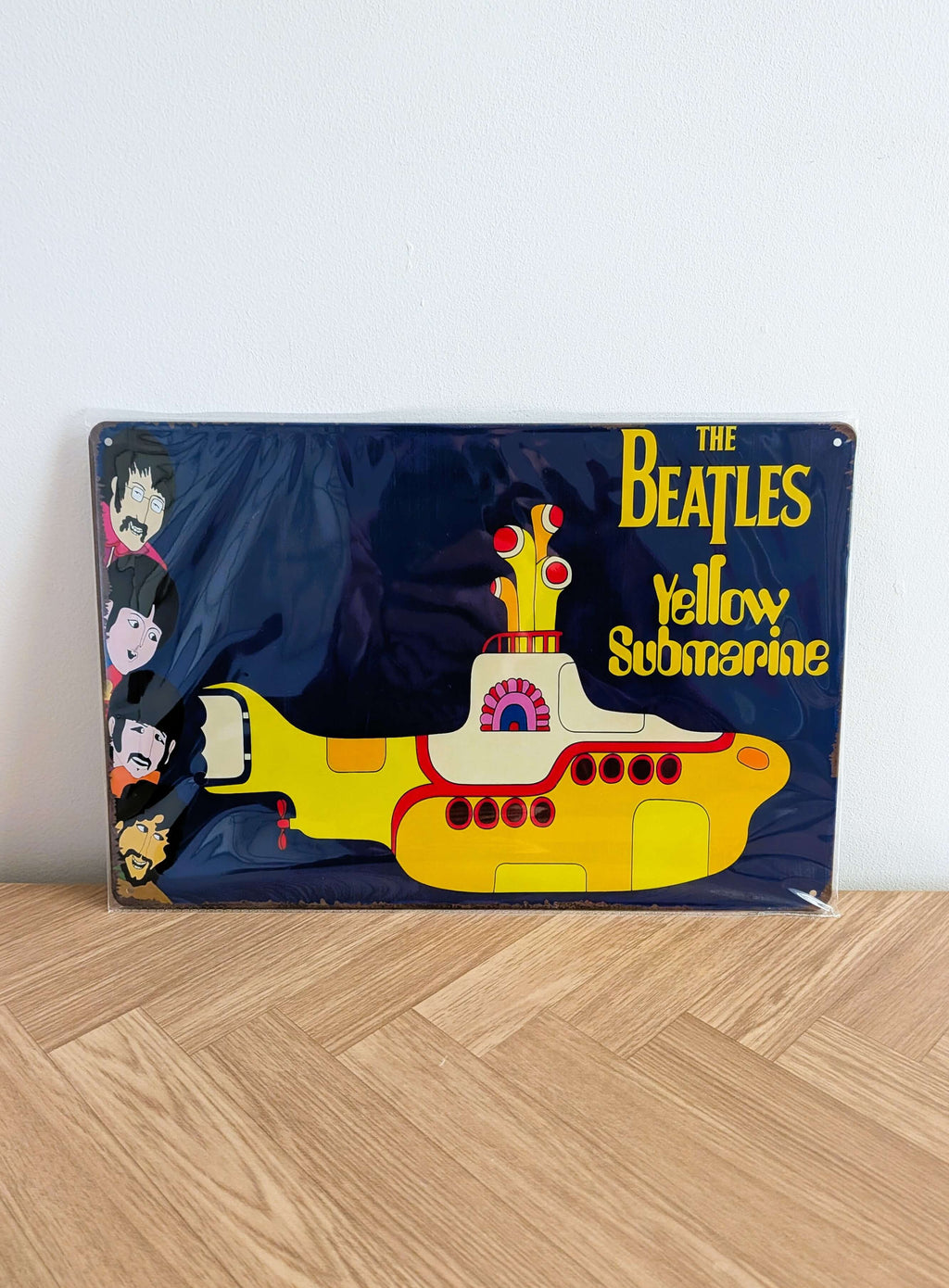 Horizontal retro-style metal wall sign for The Beatles Yellow Submarine in protective plastic wrap, featuring the iconic yellow submarine illustration, animated faces of John, Paul, George, and Ringo on the left, and bold yellow movie typography against a deep navy blue background with rusted edges.