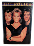 Vertical glossy metal wall sign for The Police in protective plastic wrap, featuring a classic promotional portrait photograph of Andy Summers, Sting, and Stewart Copeland against a black background, topped with a red, blue, and yellow stripe design and bold gold text.