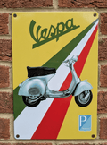 Vertical metal wall sign for Vespa featuring a classic light blue scooter over a diagonal green, white, and red Italian flag background with a yellow top section and Piaggio logo.