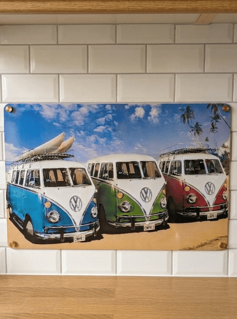 Horizontal glossy metal wall sign mounted on a white tiled wall, featuring a trio of classic vintage VW split-screen campervans in blue, green, and red parked on a sandy beach under a blue sky with palm trees.
