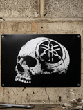 Horizontal matte black metal wall sign mounted on a concrete block wall, featuring a distressed white graphic of a human skull facing sideways with the iconic Yamaha tuning forks logo insignia stamped onto the side of the temple.